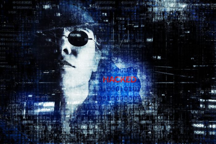 A man in dark glasses behind a computer screen with the word HACKED on it