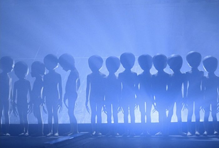 A depiction of a group of grey aliens with a bright light behind them