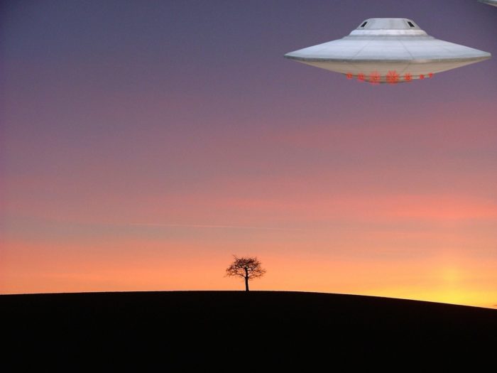 A silver UFO hovering over a lone tree at sunset