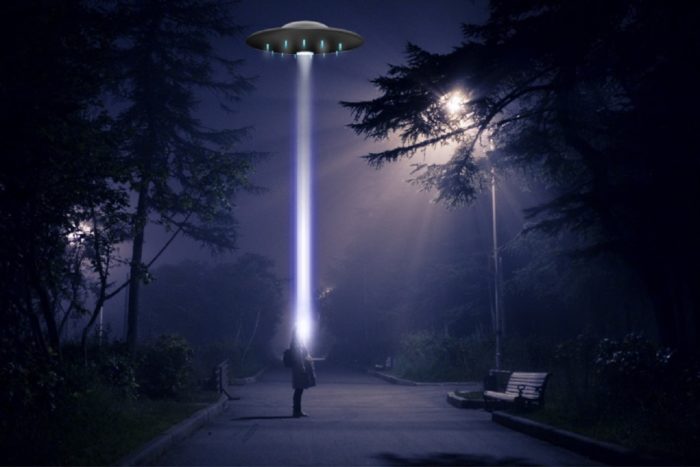 A picture of a person on a dark street looking up at a UFO with a light shining on them